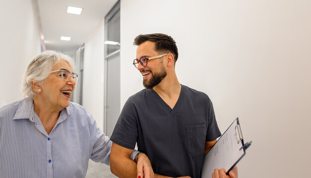 Male ophthalmologist and elderly patient laughing while discussing medical reports and walking arm in arm in hospital lobby