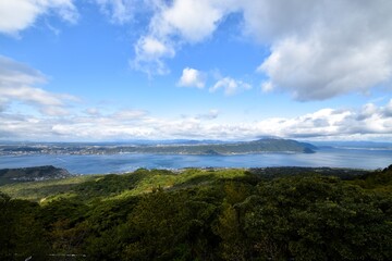 A magnificent panoramic view of Mount Sakurajima and Kinko Bay seen from Yoshino Park in Kagoshima Prefecture, Japan, featuring lush green forests and a vast blue sky with white clouds.
