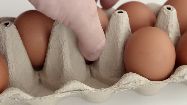 Carton of brown eggs with human hand fingers carefully selecting one egg in close-up composition emphasizing natural shell texture