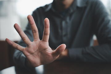 Closeup of a man's hand with fingers spread out in a stop gesture. The man is wearing a grey shirt and the background is blurred. .