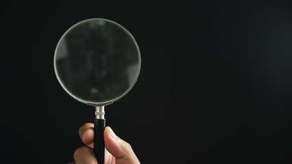 A hand holding a magnifying glass against a black background. The magnifying glass is circular and has a black handle.