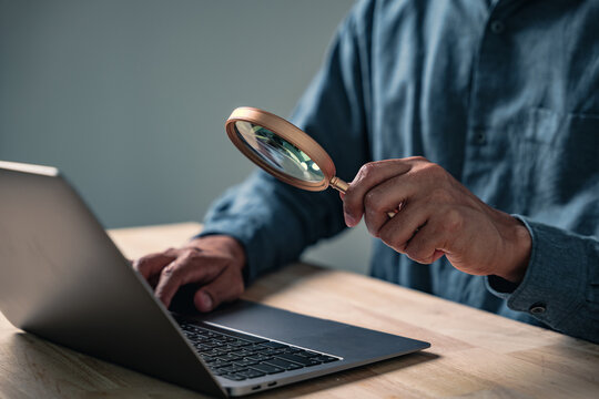 A professional man makes a hand gesture in an investigative business pose, closely examining a computer while posing with a magnifying glass for data research and analysis concepts.
