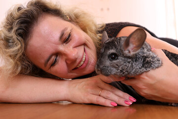 Smiling woman gently hugging a chinchilla while lying on the floor, creating a warm and trusting home scene