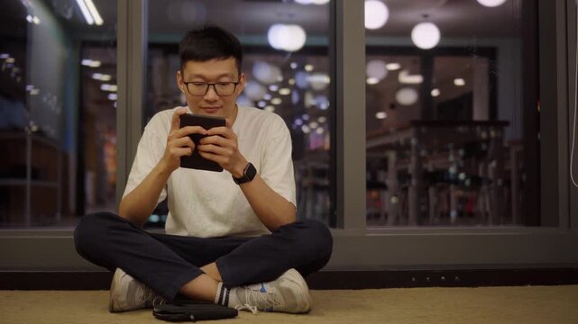 Asian man sitting on the floor and reading on an electronic device in a modern indoor space at night. The scene represents digital reading, focus, self-learning, technology lifestyle, and quiet person
