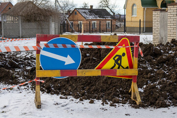 Signs indicate direction and warn of construction work on a snowy road near houses in a residential neighborhood