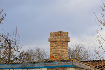 Chimney made of bricks rises above a roof with trees around it under the cloudy sky, depicting a quiet rural setting in springtime