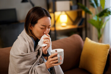 Sick Asian woman blowing nose with tissue while sitting on sofa