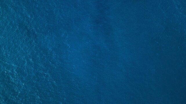 Top-down aerial drone view of deep blue sea surface with shimmering sunlight reflections and gentle wave texture, natural ocean background with copy space