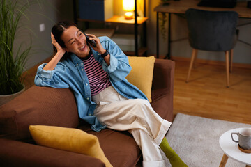 Happy Asian woman talking on smartphone while relaxing on sofa at home
