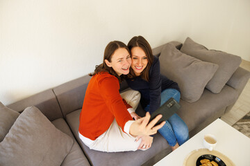 Female friends having fun taking selfies using smart phone while spending leisure time together at home
