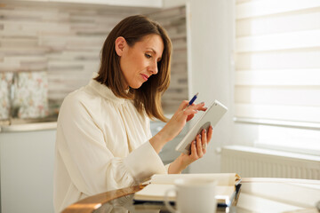 Woman writing notes in planner and using tablet computer while working remotely from home