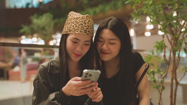 Two young asian woman gen z sisters scrolling on phone browsing at shopping mall and dating app online
