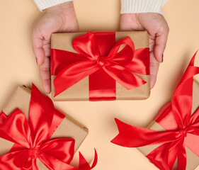 A woman's hands in a white sweater hold a gift box wrapped in craft paper with a red satin bow and a roll of ribbon. Beige background.