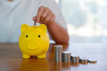 Person saving money by putting a coin into yellow piggy bank with stacked coins on table. Concept...
