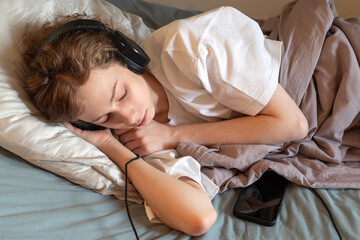Tired Teenage Boy Sleeping in Headphones with Smartphone on Bed. Authentic Concept of Digital Fatigue, Gen Z Lifestyle, Mental Health, and Relaxing Audio Content in Modern Home Interior Environment