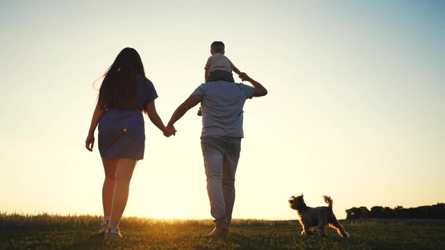 A man carries a boy family on his shoulders as a woman holds the man's hand walking on grass. The boy enjoys the view while the woman and man stroll together with their dog.