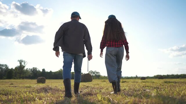 A man and woman farmers in jeans walk through a field, wearing boots as they walk on the grassy ground. The man and woman stride confidently across the open sky field.