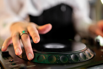 Close-up of a female DJ's hand with a ring and French manicure, manipulating a jog wheel on a professional sound mixer with vibrant green lighting in a dark nightclub. The shot captures the energy of 