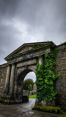 Obraz premium Ancient stone gate partly covered with green ivy and moss, overcast sky.