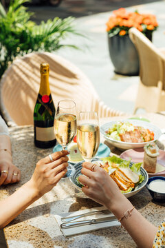 Close-up of two people clinking champagne glasses during a luxury lunch at a bright outdoor restaurant