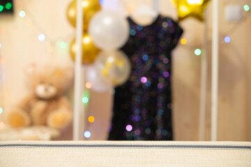 A festive blurred background with balloons, a sparkly baby dress on a hanger, and glowing fairy lights. Happy birthday concept. Focus on the foreground
