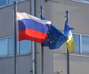 Flags of Russia, the European Union, and Ukraine wave together, blown by a strong wind. Concrete building on background.