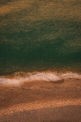 Top-down view of gentle ocean waves washing onto sandy beach with warm sunlight tones, creating a calm natural shoreline texture ideal for background, travel, wellness, and minimal design concepts.
