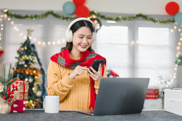 Smiling Asian woman wearing headphones and using a smartphone while sitting at a laptop in a festive Christmas home office with holiday decorations and lights.