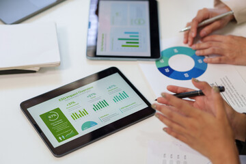 Business professionals analyzing ESG impact data and financial reports on digital tablets and printed charts during a collaborative office meeting.