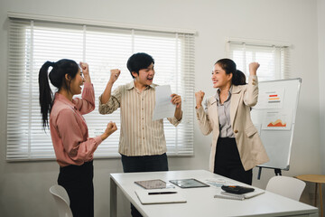Group of happy young Asian business people celebrating success and cheering with raised fists while holding document in modern office meeting room.