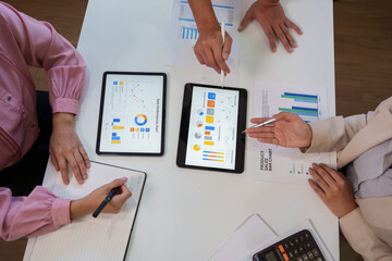 Business people collaborating on data analysis during a meeting, reviewing sales performance charts on digital tablets and printed documents in a professional office workspace.