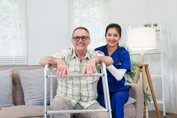 Happy elderly man using a walker while a smiling young female nurse provides professional home care assistance in a bright living room.
