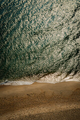 Top-down view of gentle ocean waves washing onto sandy beach with warm sunlight tones, creating a calm natural shoreline texture ideal for background, travel, wellness, and minimal design concepts.