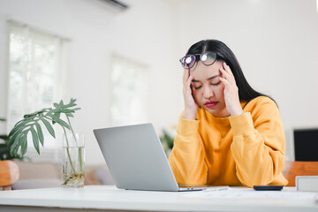 Stressed young Asian woman holding her head with closed eyes, suffering from a headache or exhaustion while working on a laptop in a bright home office.