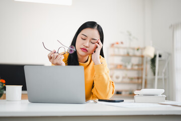 Tired young Asian woman holding her glasses and closing her eyes while suffering from stress and fatigue at her home office laptop desk.