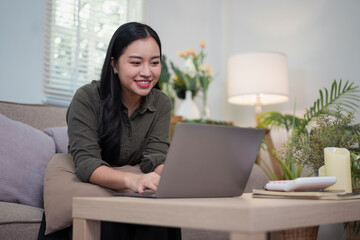 Happy young Asian woman smiling while working on a laptop computer on a coffee table in her living room at home.