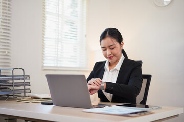 Young Asian businesswoman checking the time on her wristwatch while sitting at a desk with a laptop in a bright professional office.