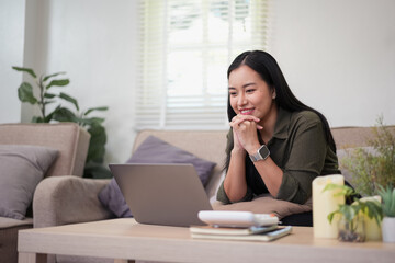 Smiling Asian woman working on a laptop and using a calculator while sitting on a sofa in a bright living room at home.