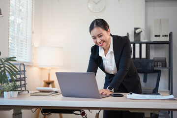 Smiling Asian business woman in a black suit standing at a desk and using a laptop while working in a bright modern home office.
