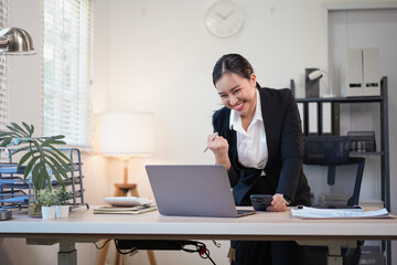 Happy Asian business woman celebrating success and cheering while looking at laptop and holding smartphone in a bright office workspace.