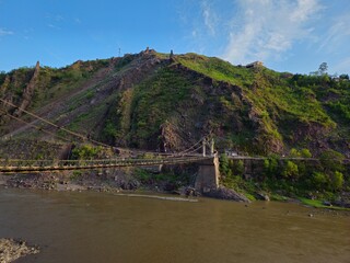 A historic suspension bridge spans a wide river at the base of a lush, rocky mountain under a clear blue sky.