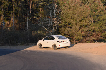 White sedan parked beside a curved mountain road surrounded by forest. Car stopped along a scenic hillside road in a natural landscape. Travel scene with a white car on a winding forest road.