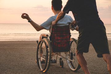 Rear view of a person with disability in a wheelchair being pushed by his friend or caregiver on the sandy beach during golden hour. Support and outdoor activity for people with limited mobility.