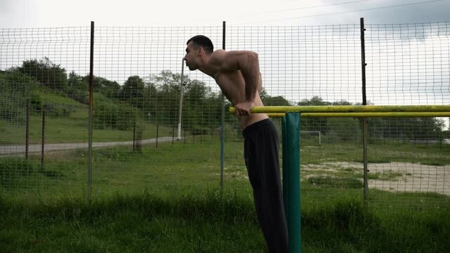 Young sportsman doing push ups on parallel bars at nature. Strong sporty man exercising at sports ground. Male shirtless athlete training hard outdoor. Concept of active sportive lifestyle. Slow mo