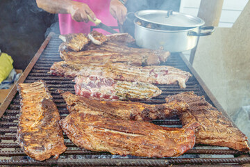 A rack of ribs of meat being grilled over hot coals, while a cook handles it with metal utensils.