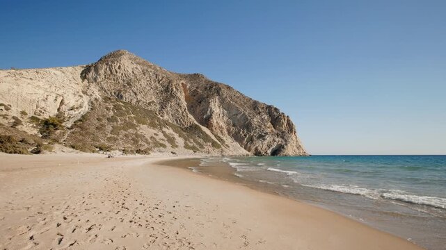 Magical empty sandy beach on the Greek island of Kos, washed by the waves of the clear blue sea on a sunny day with a rocky mountain in the background