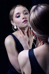 Portrait of young blonde woman near mirror. Female in black dress with straps and earring.