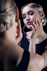 Portrait of young blonde woman near mirror. Female in black dress with straps and earring.