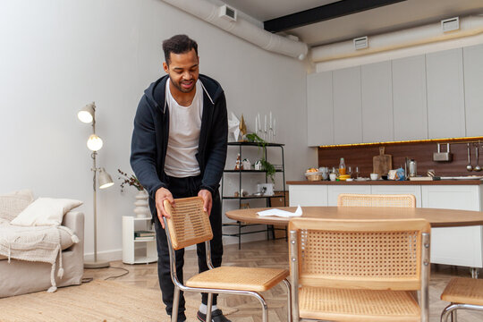 African American man cleans his house and puts wooden chair under the table, the guy carries furniture and smiles