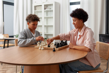African American woman and her son play chess at home at table, curly-haired teenager and his mother compete in educational board game